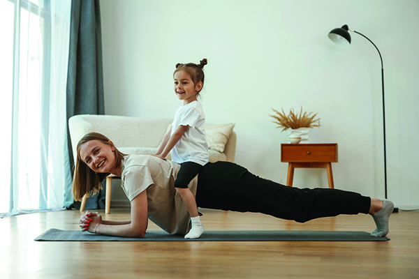 Kid is sitting on a back of woman. Young mother with little girl are doing yoga at home.